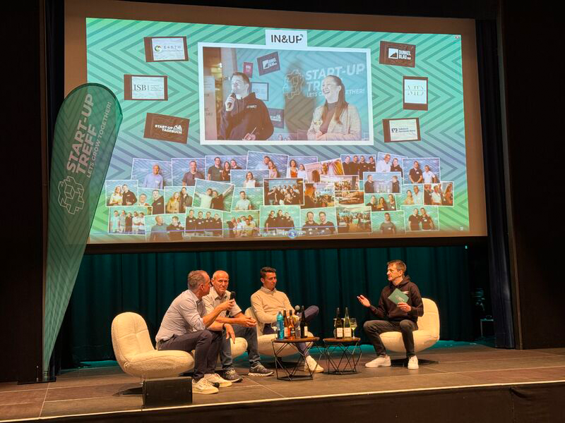 Nahaufnahme des Podiums mit 3 Personen, die miteinander reden. Im Hintergrund eine Leinwand mit einer aktiven Folie.