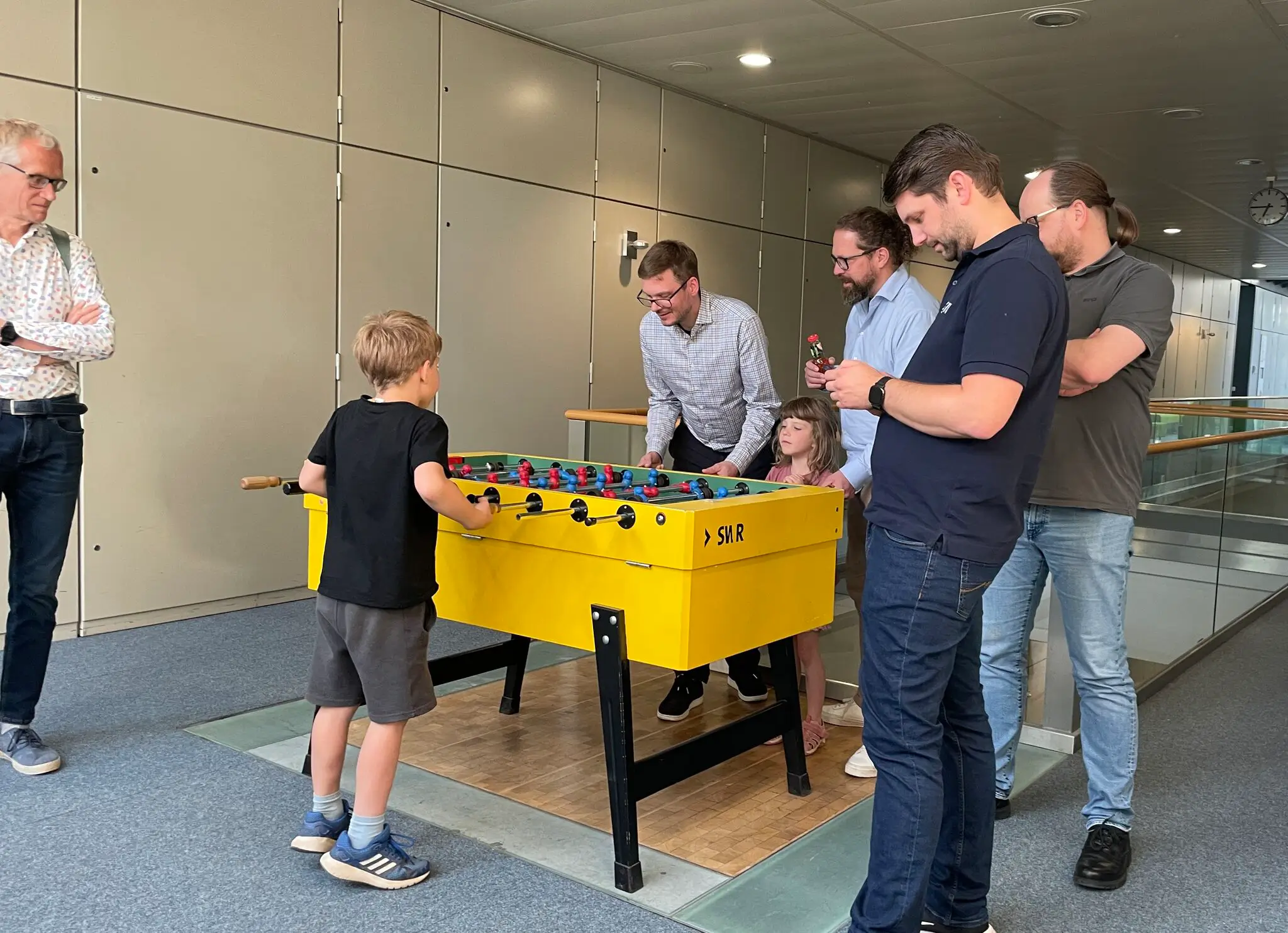 Fünf Erwachsene und zwei Kinder versammeln sich um einen Kickertisch und spielen mit Begeisterung beziehungsweise schauen zu